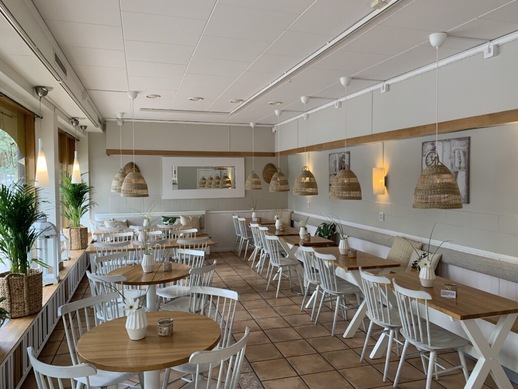 Cafédelen hos Ritz Konditori med ljusa träbord, vita pinnstolar, rottinglampor och gröna växter.